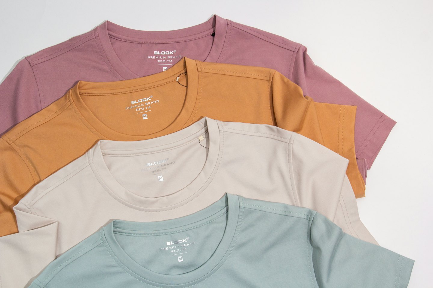 Cuatro camisetas dobladas en rosa empolvado, naranja quemado, beige y verde salvia.